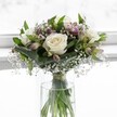 An elegant white rose bouquet stands poised in a clear cylindrical glass vase, set before a softly glowing white background that suggests a bright Oxford morning filtering through a window. Three fully opened, creamy white roses form the calm, luxurious heart of the arrangement, each bloom with gently unfurling petals that spiral inward to a faint ivory centre, giving an impression of purity and quiet confidence. Around them, glossy green foliage fans out in layered arcs, with some broad leaves carefully folded to cradle the flowers, creating a structured, rounded shape that feels deliberate and expertly composed. Threaded through the greenery are delicate sprigs of white gypsophila, or baby's breath, their tiny star-like flowers floating in airy clusters, like a light mist above Port Meadow at first light. Slender stems of alstroemeria, some in bud and some softly opening, introduce subtle tones of muted mauve and pale blush-pink, adding gentle colour without disturbing the bouquet's serene, almost monochrome calm. The stems are visible through the crystal-clear water in the vase, gathered neatly and secured just above the waterline with a fine, barely noticeable ribbon. The light catches the water and glass, echoing the soft luminosity of the roses and enhancing every curve and texture. The overall mood is tranquil and contemplative, perfect for a thoughtful gesture of sympathy, a peaceful wedding bouquet at an Oxford college chapel, or a quiet thank-you delivered to a townhouse off the High Street. Created with the sensitivity of a local florist, the arrangement radiates stillness, grace, and a sense of gentle, enduring care.