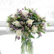 A softly lit sympathy bouquet stands tall in a transparent glass cylinder vase, its reflection subtly catching the light as though positioned near a bright Oxford window overlooking a calm side street off Banbury Road. The flowers are arranged with deliberate care, dominated by creamy white roses whose petals curl open in flawless, rounded layers. These roses are interlaced with stems of alstroemeria showing gentle shades of purple, from pale lavender to deeper mauve, with touches of fresh green at the buds, adding a muted whisper of colour without breaking the overall serenity. Throughout the bouquet, delicate clusters of white baby's breath form tiny, cloud-like puffs that bridge the spaces between the larger blooms, giving the design an ethereal, almost weightless quality. The foliage is a deep, polished green, with broad leaves and finer, tapering stems that frame and support the flowers, lending the arrangement structure and a sense of natural abundance. Inside the clear vase, the straight green stems are visible through the clean water, their vertical lines adding a quiet order that suits the reflective mood. The background is bright and softly blurred, nearly pure white, suggesting a peaceful interior, perhaps a calm sitting room in central Oxford or a quiet study in Jericho, where the bouquet can be a gentle, steady presence. The overall feel is one of comfort and dignity, crafted as a tender expression of sympathy and remembrance, appropriate for sending to a grieving family or accompanying a memorial gathering with understated grace.