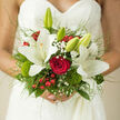A hand-tied bridal bouquet is cradled against the delicate lace of a wedding gown, its design a beautiful contrast of white lilies and passionate red roses that feels perfectly at home in an elegant Oxford celebration. The lilies, creamy and star-shaped, open generously toward the light, their petals curving in soft, fluent lines that suggest purity and calm. At the heart of the bouquet, a rich red rose anchors the composition, its velvety petals spiralling inward like a secret promise spoken in the cloisters near the Bodleian Library or along Broad Street. Around these central blooms, airy sprigs of baby's breath drift like fine mist, their tiny white flowers filling the spaces with tenderness. Glossy red berries and bright moss-green dianthus add joyful pops of colour and playful texture, reminiscent of the vibrant greenery in University Parks after a spring shower. The surrounding foliage is lush and varied in tone, from deep forest greens to fresher lime shades, each leaf catching hints of light and shadow. Slender blades of grass and trailing greens skim the edges, giving the bouquet a slightly wild, romantic character rather than something too formal or stiff. The lace of the dress forms a soft backdrop, its intricate pattern echoing the careful, thoughtful way the bouquet has been arranged. The whole scene evokes the quiet emotion just before a ceremony begins in one of Oxford's historic venues, a bouquet that seems to hold not only fragrance and colour, but the anticipation, joy, and devotion of saying I do in this storied city.