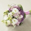 Resting upon a soft, neutral-toned, lightly textured surface that calls to mind the tranquil charm of Oxford's riverside settings, this hand-tied bridal bouquet captures the essence of contemporary elegance with a local twist. At its heart are sumptuous white lisianthus flowers, their ruffled petals layered exquisitely, each pale center softly tinged with creamy yellow. Unopened buds nestle beside pastel accents of blush pink and lavender stock, their frothy, diminutive blossoms bringing gentle movement and a touch of romance, reminiscent of blossoms seen in Oxford's private courtyard gardens. Interwoven rich green foliage and bright lisianthus buds add lively structure, while the striking deep purple heads of Allium 'Star of Persia' punctuate the softness, their spiky globes creating bold visual intrigue much like the surprise of wildflowers on a walk through Headington Hill Park. The bouquet is rounded, slightly elongated, and hand-tied with a radiant fuchsia pink satin ribbon, finished with an elegant winding of white pearls-a subtle tribute to Oxford's enduring love of tradition. Soft, even lighting reveals each petal's delicate translucency and the careful florist's artistry behind every detail. This bouquet is perfectly suited for a romantic Oxford ceremony near Carfax Tower or as the centerpiece of a joyful garden celebration, its design reflecting both contemporary wedding trends and the timeless beauty cherished in the heart of the city.