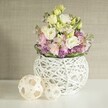 A softly lit still life features a rounded white vase on a bright, clean tabletop, overflowing with pastel blooms that seem to glow against a gently textured greige wall. The vase itself is a modern sphere made from an intricate open weave of white rattan or painted twigs, its airy gaps giving glimpses of the inner container and adding a crafted, almost sculptural quality often seen in boutique interiors around Oxford city centre. From this nest-like form rises a generous bouquet of creamy white and pale yellow eustoma, or lisianthus, their many ruffled petals unfolding like little clouds. Threaded between them are slender stems of soft pink stock flowers, their clustered florets climbing upwards to give a vertical rhythm and a sense of lush movement, as though the bouquet is stretching towards soft morning light over the Cherwell. At the base, tiny vivid purple filler blooms, reminiscent of asters or small alliums, create a rich, textural carpet that deepens the colour palette and makes the pale blossoms appear even more luminous. A simple collar of deep green foliage encircles the arrangement, grounding the pastels with fresh, natural contrast. To the left of the main display, two decorative spheres in lighter natural wood or rattan echo the vase's open-work pattern, reinforcing the theme of handwoven textures and calm, organic design you might find in a quiet Jericho townhouse. The overall mood is serene and elegant, conjuring the feeling of a peaceful spring morning and making this arrangement ideal for a refined dining table, a thoughtful anniversary gift, or a welcoming hallway display in an Oxford home.