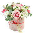 A graceful floral hatbox arrangement is showcased against a pristine white background, its soft pastel tones and careful craftsmanship creating an impression of quiet luxury that would feel at home in any Oxford drawing room. The container is a round, blush-pink hatbox with a smooth, matte surface, its gentle colour echoing the first flush of blossom along the River Thames in spring. Encircling the box is a narrow coral-red satin ribbon, neatly tied into a small bow at the front; from this bow hangs a cream tag decorated with green script, ready to carry a personal message for a birthday in Summertown or an anniversary in central Oxford. Emerging from the top, the bouquet rises in a generous, domed shape, each stem placed with intention. At the centre, full-petalled pink roses unfurl in layered spirals, some shaded with deeper pink or fuchsia at their core, drawing the eye with a romantic, focal glow. Around them, clusters of white daisy-like chrysanthemums with fresh green centres provide a crisp contrast, their simple shapes adding a light-hearted, uplifting energy. White alstroemeria, identifiable by their slender trumpets and fine inner speckles, weave gracefully through the design, while smaller deep pink flowers enliven the arrangement with brighter notes of colour. Eucalyptus leaves in soft, silvery-green tones arc between the blooms, their rounded, slightly waxy surfaces catching the light and lending a contemporary, airy structure. Deeper green foliage and a few delicate twigs tipped with tiny buds or seed pods add subtle texture and natural charm, giving the bouquet a just-arranged freshness. The overall impression is one of tenderness and celebration, ideal for marking special moments or sending a thoughtful gesture anywhere across Oxford.