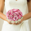 Cradled tenderly against an intricate lace wedding dress, this bridal bouquet of blush pink roses and airy white gypsophila appears as if it were painted in soft watercolours across a calm Oxford afternoon. Each rose is fully open but still fresh, its petals layered in rounded, silky spirals that create a gentle dome of pink, warmed at the centre and fading to pale at the outer edges. Clusters of baby's breath nestle between the blooms, hundreds of tiny white flowers forming a delicate mist that softens the lines of the bouquet, much like morning fog drifting over Port Meadow. The design is carefully composed and evenly proportioned, with no single rose overpowering the others, giving the impression of balance and quiet confidence. You can almost sense the coolness of the stems wrapped together beneath her fingers and the soft, powdery fragrance of newly opened roses mingling with the faint, grassy freshness of the gypsophila. The colours remain true to a romantic palette-muted pinks, creamy whites, and hints of tender green-made richer by the gentle, natural light falling across the scene, as though the bride were standing near a tall window overlooking Oxford's historic rooftops. The bouquet's rounded shape sits gracefully in her hands, aligned with the lines of her dress so that lace, flowers, and skin form one harmonious composition. Emotionally, it suggests tenderness and heartfelt devotion rather than grandeur: the kind of bouquet chosen by someone who values sincerity, tradition, and quiet elegance. Every bloom seems to echo the words of a lifelong promise, ready to accompany her from the first step down the aisle to the last dance of the evening.